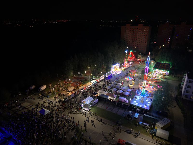 Szeroki kadr terenu imprezy plenerowej nocą, karuzele i bloki w tle